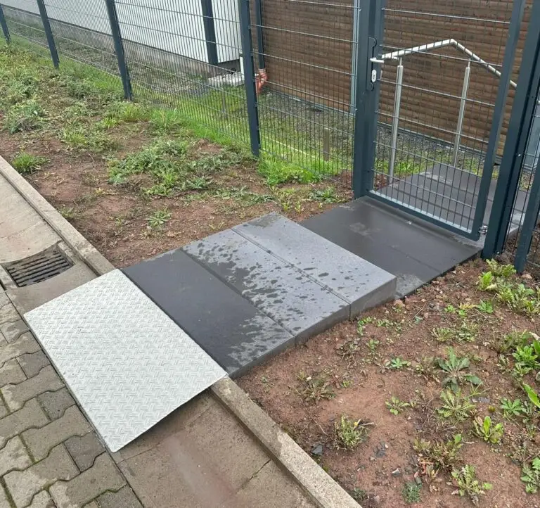 Metallrampe über einen Bordstein zum Eingangstor einer eingezäunten Anlage